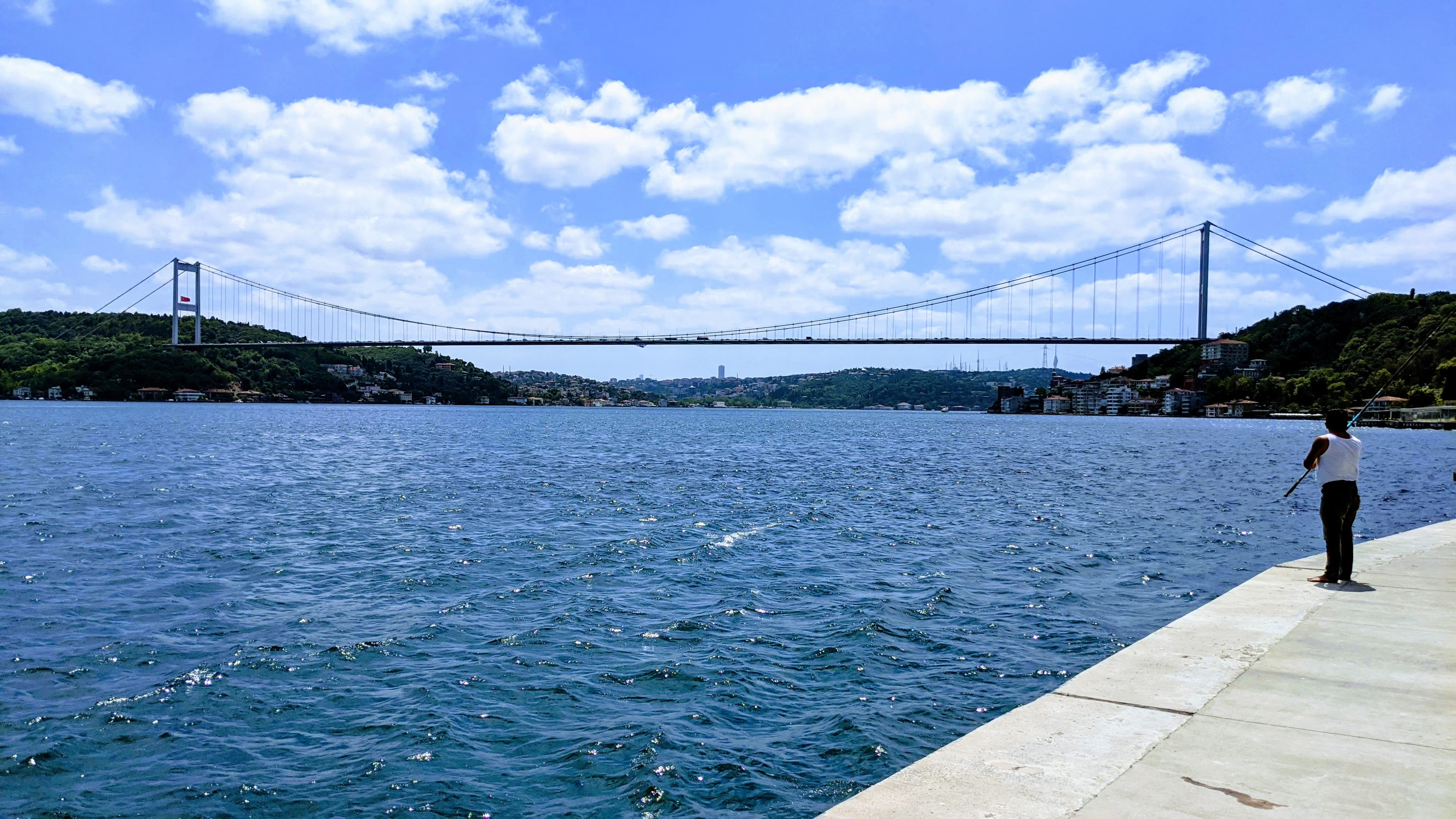 Fisherman on the Bosphorus shore with the bridge spanning two continents behind him, Istanbul